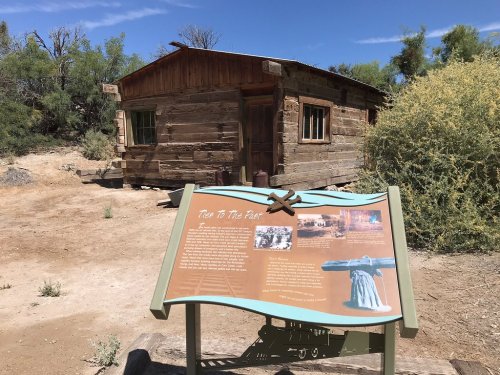 A rustic wooden cabin with a historical sign in front, surrounded by sparse vegetation and dry ground.
