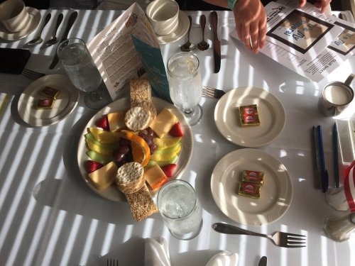 A table set with plates of fruit, cheese, crackers, and butter, surrounded by glasses and utensils, with shadows from blinds.