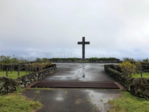 A large cross stands on a hilltop, surrounded by greenery and a cloudy sky, with a pathway leading up to it.