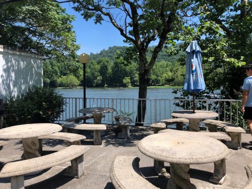 A scenic view of a river with stone tables and chairs under trees, featuring a blue umbrella in the background.