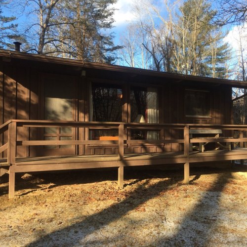 A wooden cabin with a porch, surrounded by trees and a clear blue sky. The ground is partially covered in snow.