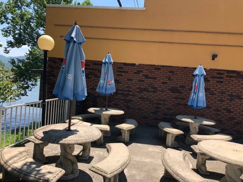 Outdoor seating area with stone tables and blue umbrellas, overlooking a body of water and surrounded by greenery.