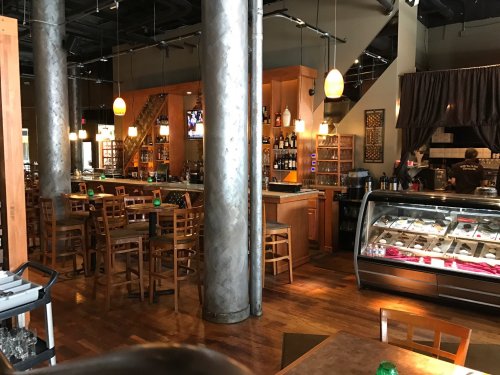 Interior of a modern restaurant featuring a bar, wooden seating, and a display case with desserts. Warm lighting throughout.