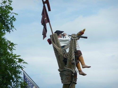 A pirate figure with a parrot sits atop a wooden lookout post, holding a telescope against a cloudy sky.