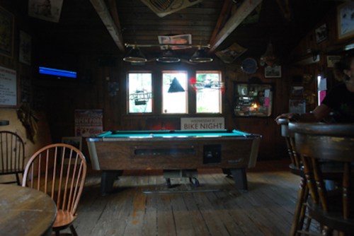 A rustic bar interior featuring a pool table, wooden chairs, and dim lighting, with a sign for "Bike Night" in the window.