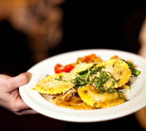 A hand holding a plate of ravioli topped with herbs and served with colorful vegetables.
