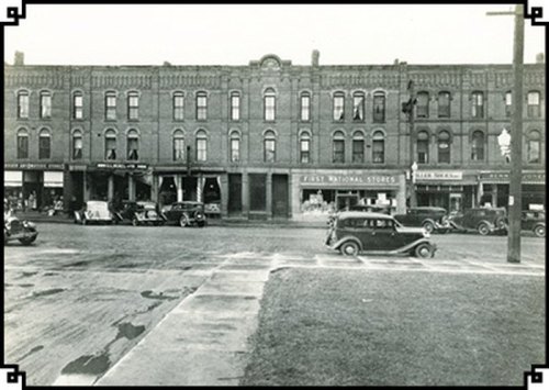Historic black-and-white photo of a street scene with vintage cars and early 20th-century storefronts.