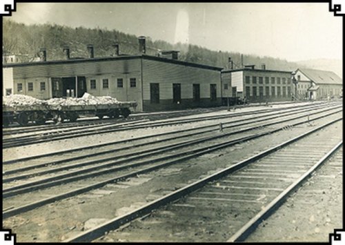Black and white photo of a railway yard with tracks, buildings, and a freight car loaded with materials.