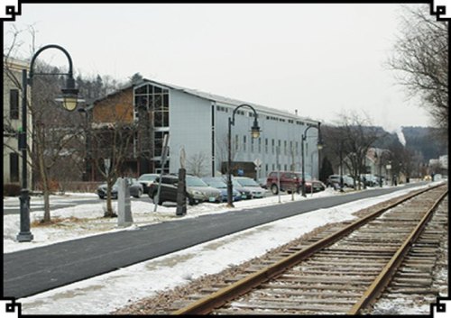 A snowy street scene with a railway track, parked cars, and buildings in the background under a gray sky.