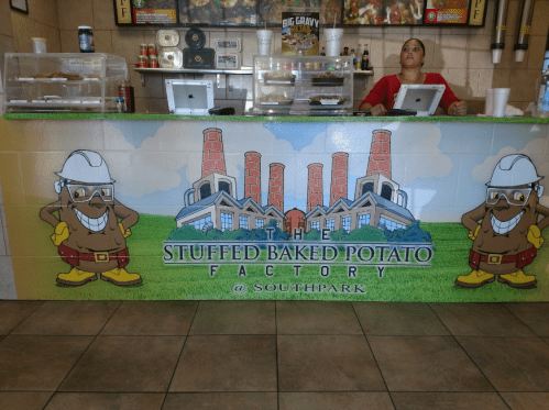 A colorful mural of a cartoon character in hard hat at the Stuffed Baked Potato Factory counter with a staff member behind.