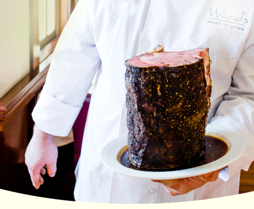 A chef in a white coat holds a large, beautifully cooked prime rib on a plate, with a rich sauce underneath.
