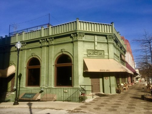 A green historic building with large windows and an awning, located on a sunny street corner.