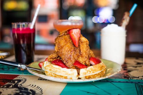 A plate of fried chicken on waffles, topped with strawberries, alongside colorful drinks in glasses.