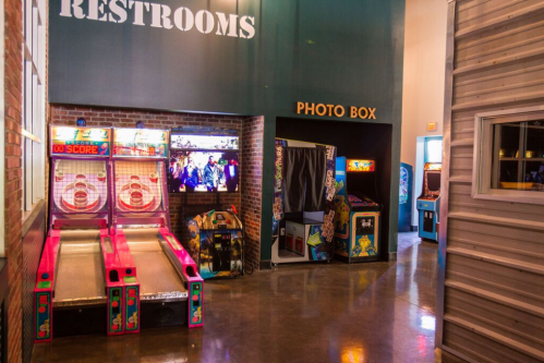 A vibrant arcade area featuring skee-ball machines, a photo booth, and various arcade games under a "Restrooms" sign.