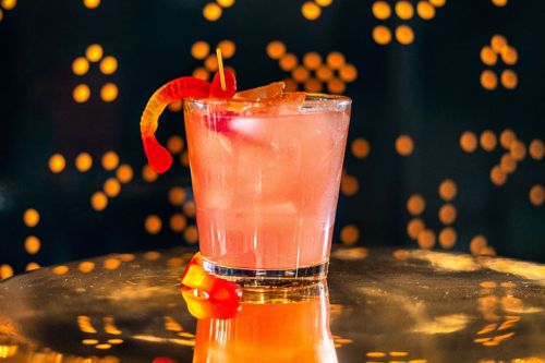 A vibrant pink cocktail garnished with a gummy worm, sitting on a reflective surface with a bokeh background.