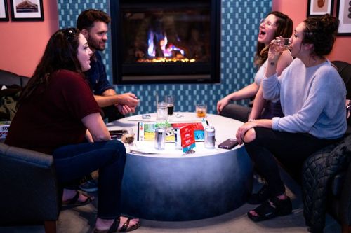 A group of four friends enjoying drinks and laughter around a coffee table by a cozy fireplace.