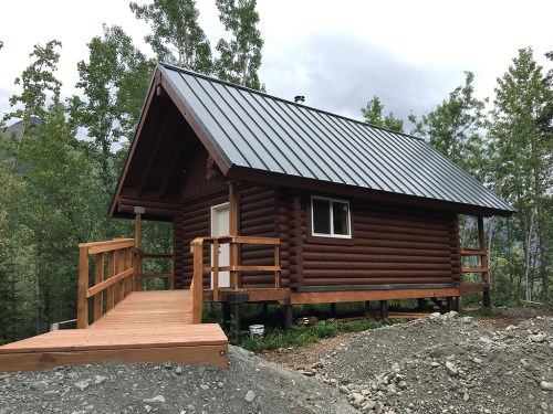 A small log cabin with a metal roof, surrounded by trees and gravel, featuring a wooden deck and steps.