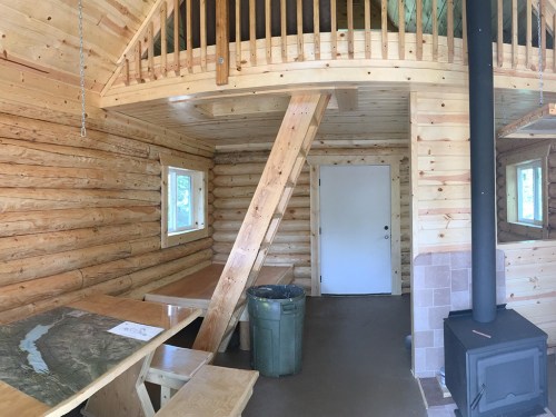 Interior of a log cabin featuring wooden beams, a loft, a table, a trash can, and a wood stove.