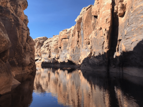 A serene canyon with towering rock formations reflecting in calm water under a clear blue sky.