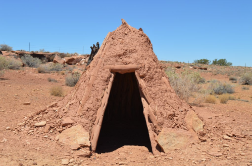 A small adobe structure with a triangular entrance, set in a dry, desert landscape with sparse vegetation.