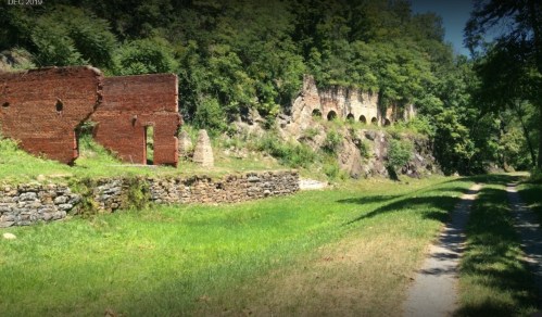 Ruins of brick structures along a grassy path, surrounded by trees and rocky terrain.