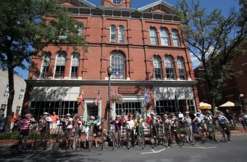 A group of cyclists with vintage bicycles poses in front of a historic brick building on a sunny day.