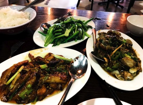 A table with three dishes: stir-fried greens, clams with vegetables, and a meat dish, alongside bowls of rice.
