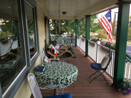 A cozy porch with a table, chairs, and an American flag, surrounded by greenery and colorful flowers.