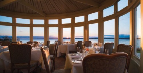 Elegant dining room with large windows overlooking a sunset by the water, featuring tables set for a meal.
