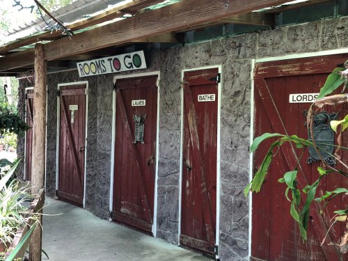 Four wooden doors with signs labeled "Rooms to Go," "Ladies," "Bathe," and "Lords," set against a stone wall.
