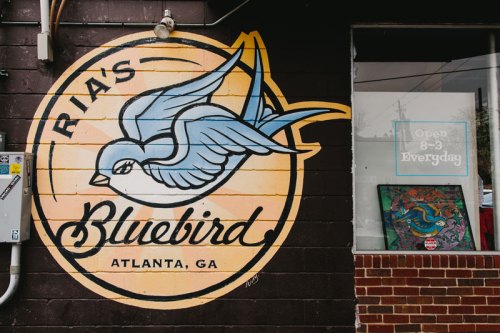 Mural of a bluebird with "Ria's Bluebird" and "Atlanta, GA" on a wall, next to a window displaying art.