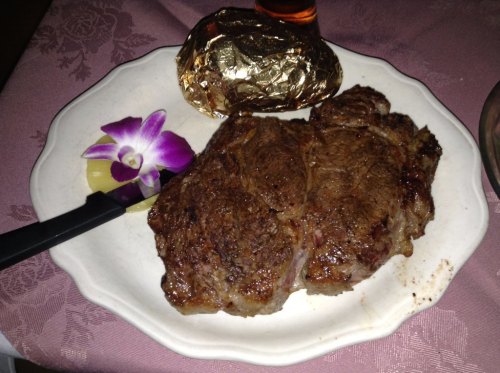 A plate with a large, grilled steak, a baked potato wrapped in foil, and a purple orchid garnish.