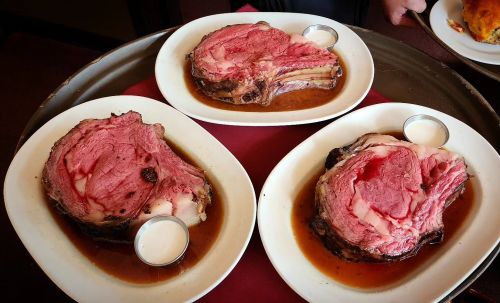 Three large servings of prime rib on white plates, served with a side of dipping sauce, on a tray with a red cloth.