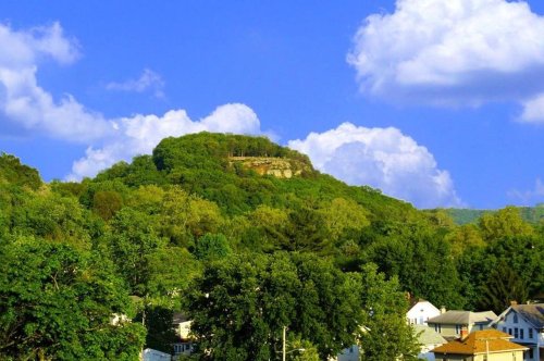 A lush green hill topped with trees under a bright blue sky with fluffy white clouds, overlooking a small town.