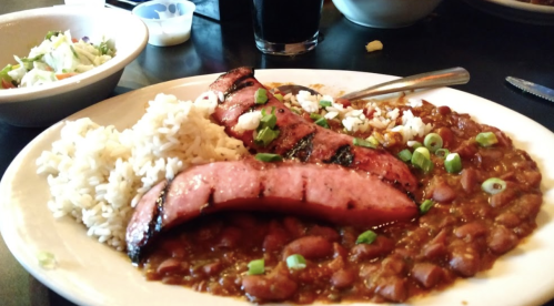 A plate of grilled sausage slices served with rice and beans, garnished with green onions, alongside a salad and drink.