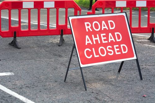 A bright red sign reading "ROAD AHEAD CLOSED" in front of red barrier fencing on a paved road.