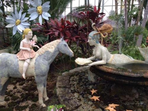 A whimsical scene featuring a girl on a horse interacting with a mermaid by a fountain, surrounded by lush plants.