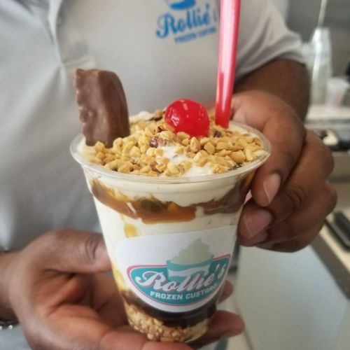 A person holds a cup of frozen custard topped with nuts, a cherry, and a chocolate treat, with a red straw.