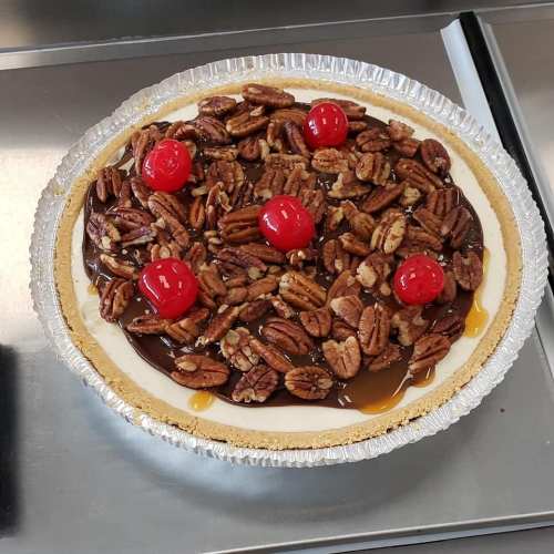 A pie topped with pecans, cherries, and drizzled caramel, resting in a silver pie dish on a stainless steel surface.