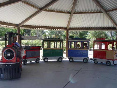 A colorful miniature train with children riding inside, under a shaded pavilion in a park.