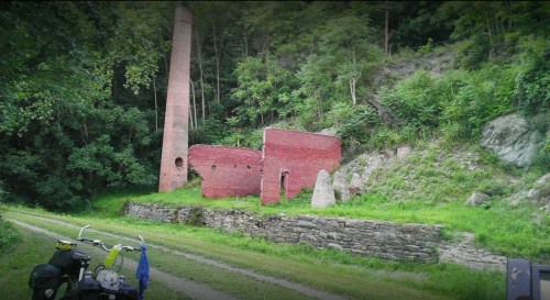 Ruins of a red brick structure with a tall chimney, surrounded by greenery along a dirt path.