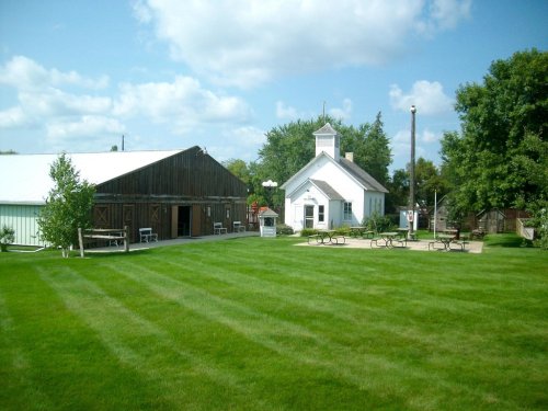 A grassy area with a barn and a small church, surrounded by trees and picnic tables under a blue sky with clouds.