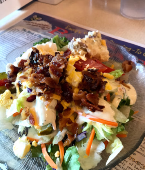 A colorful salad topped with bacon, cheese, and dressing, served on a glass plate with a side of chicken salad.