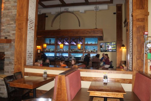 Interior of a restaurant featuring wooden decor, a bar with illuminated shelves, and patrons seated at tables.