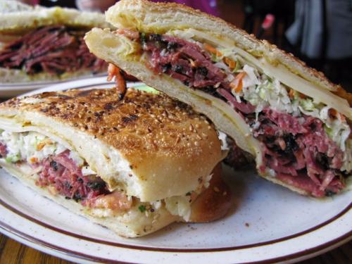 A close-up of a sesame-seed sandwich filled with layers of meat and coleslaw, served on a plate.