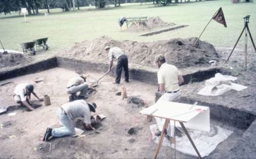 Archaeologists excavate a site, using tools and measuring equipment, with mounds of dirt and a flag in the background.