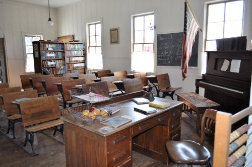 A vintage classroom with wooden desks, a chalkboard, bookshelves, and a piano, featuring an American flag.