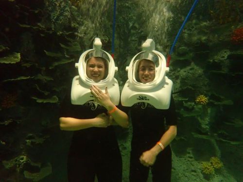 Two people in underwater helmets smile while submerged in a colorful coral reef environment.