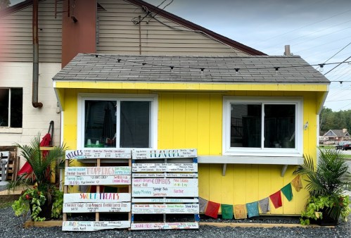 A bright yellow food stand with a menu board displaying various toppings and sweet treats, surrounded by greenery.