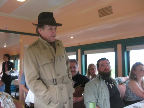 A man in a trench coat and hat stands in a dining car, while others sit at tables, some looking at him.